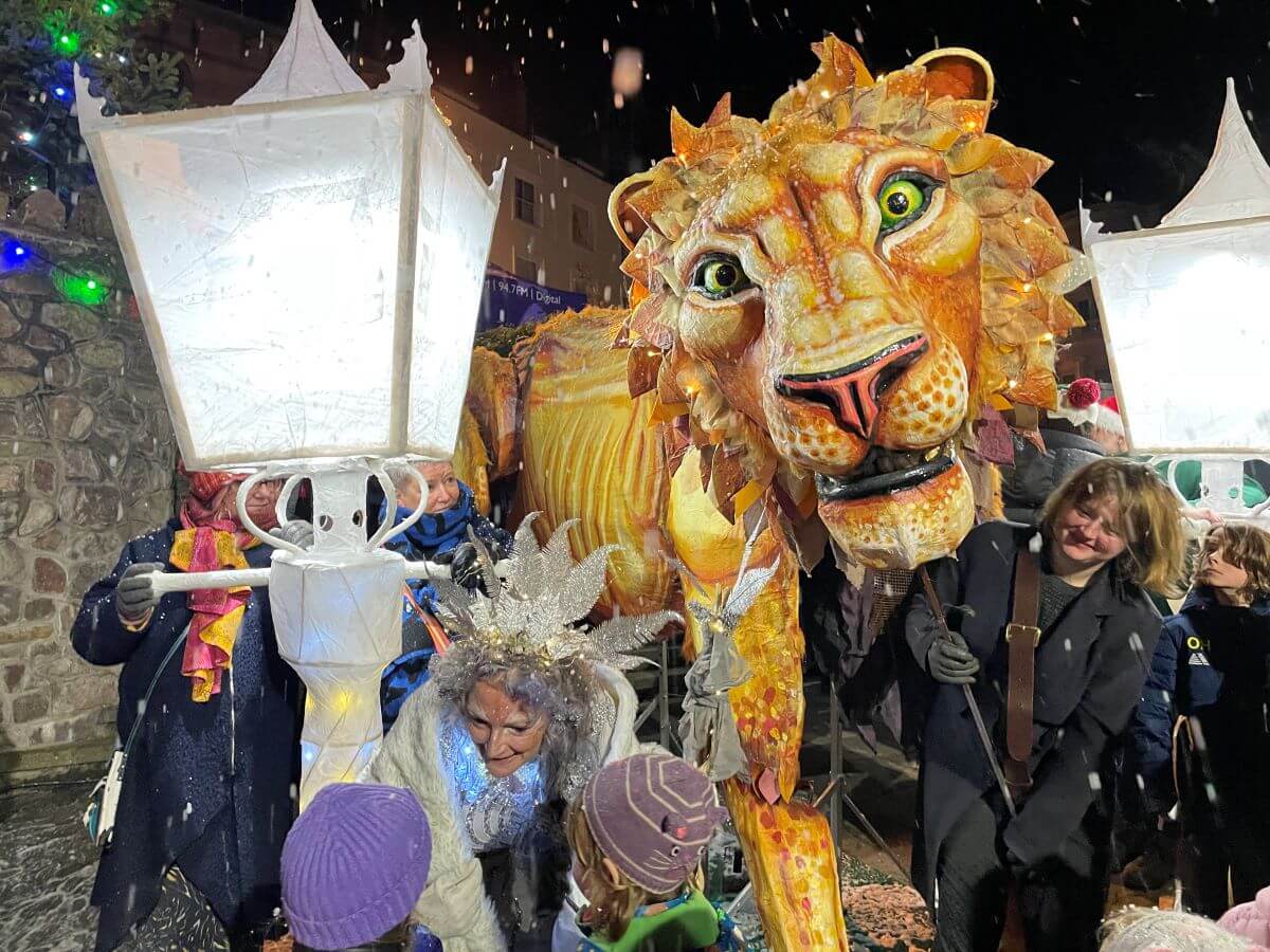 a large lion puppet in a crowd with artificial snow falling 