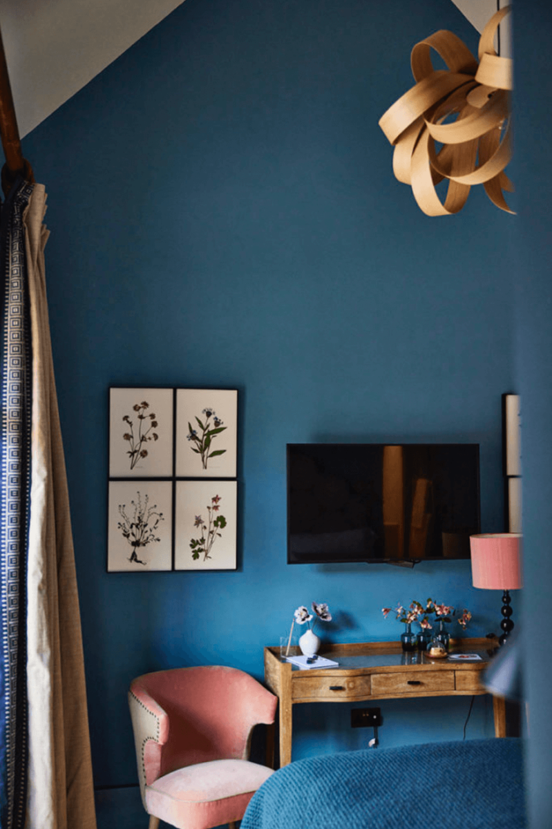 A blue painted hotel room with floral art, a pink chair and tv on the wall