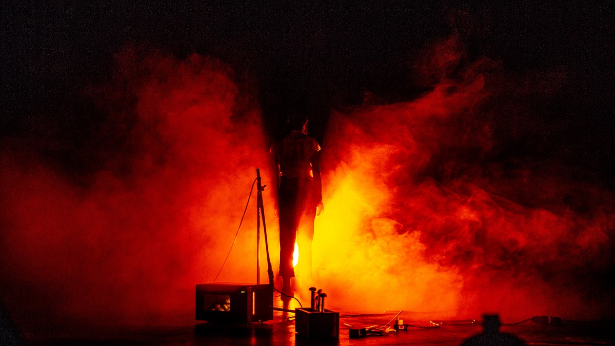 A figure stands largely in darkness, with a yellow-orange light shining towards them