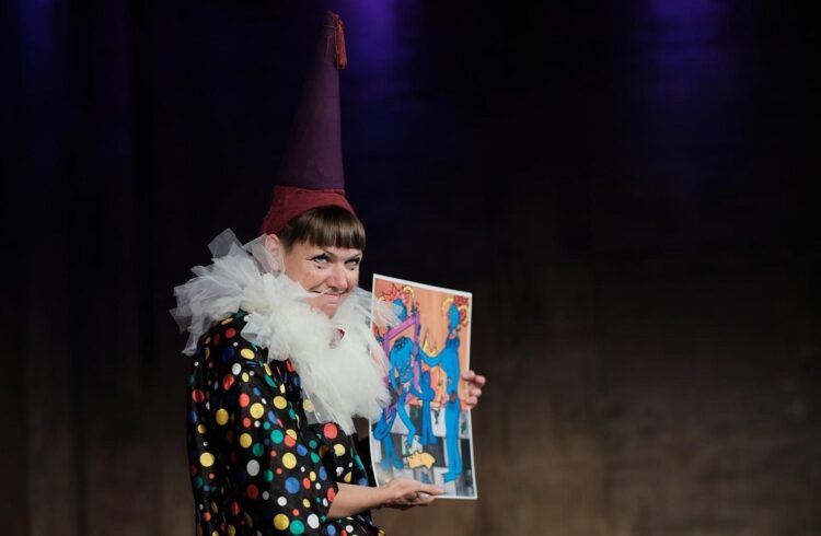 A clown on stage wearing a large hat and holding a tarot card