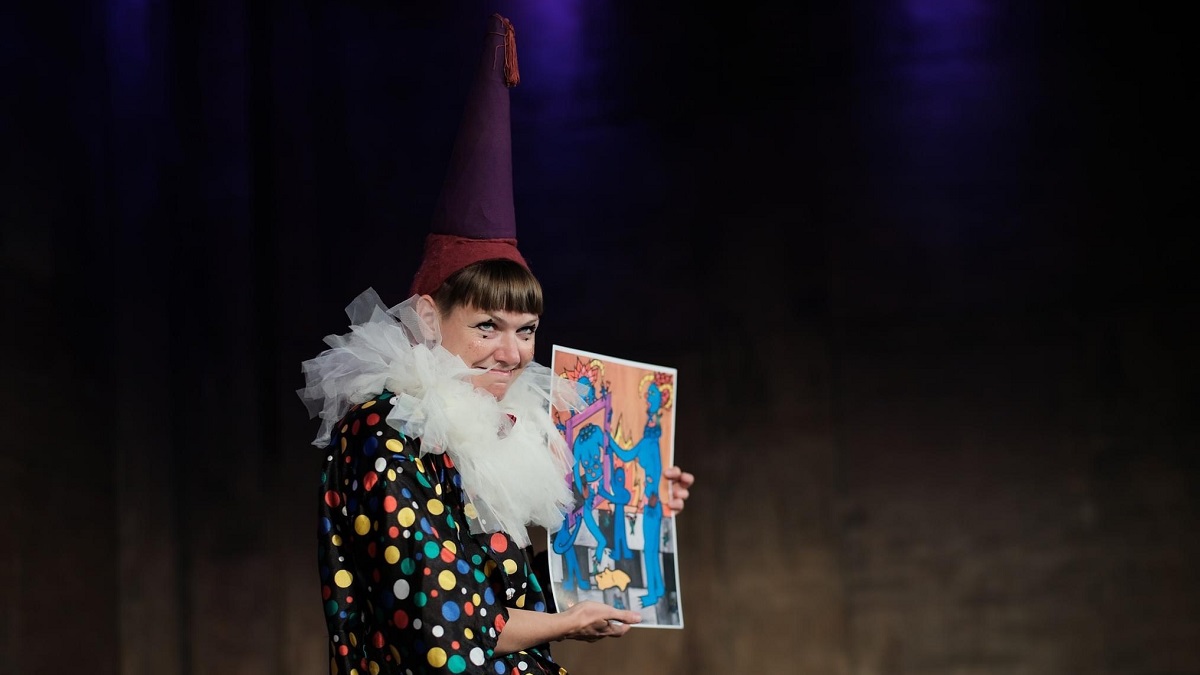 A clown on stage wearing a large hat and holding a tarot card